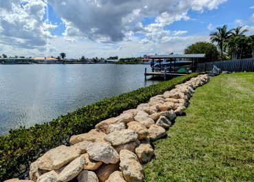 A picture after Coquina Seawall Installation on a canal with green grass and red flowers and a calm river behind