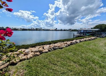 A picture after Coquina Seawall Installation on a canal with green grass and red flowers and a calm river behind